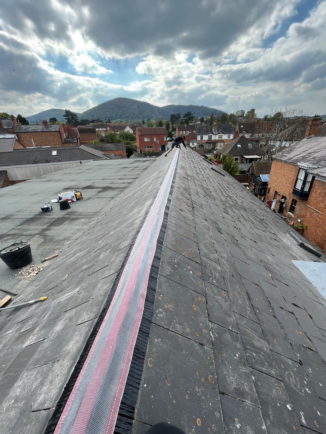 Roofer laying ventilated ridge roll along a slate roof, with hills in the distance